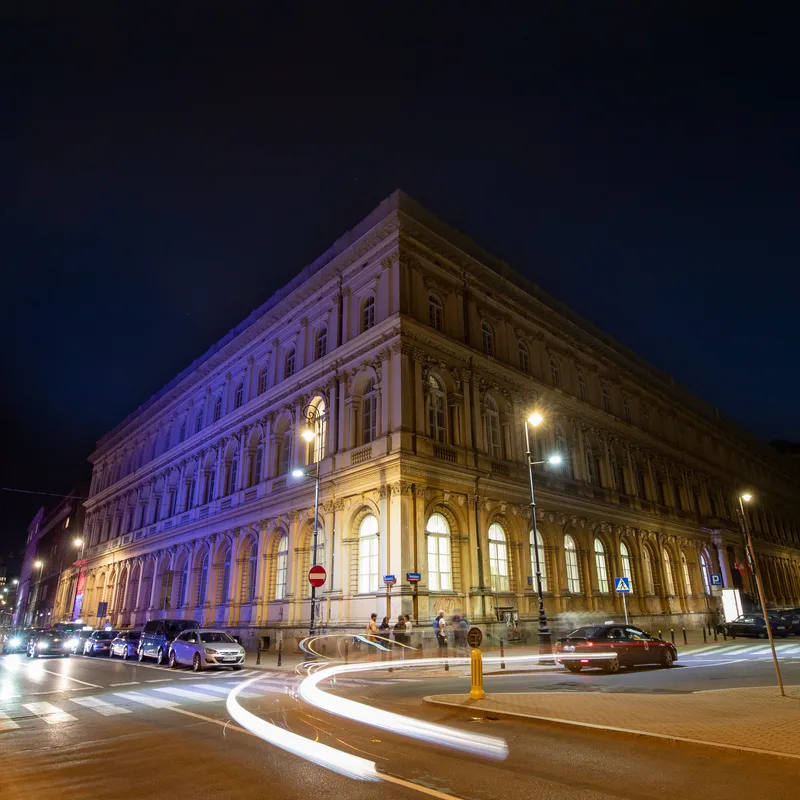 Gmach Muzeum Etnograficznego noca - neorenesansowy budynek podswietlony na fioletowo z pasami swiatla samochod